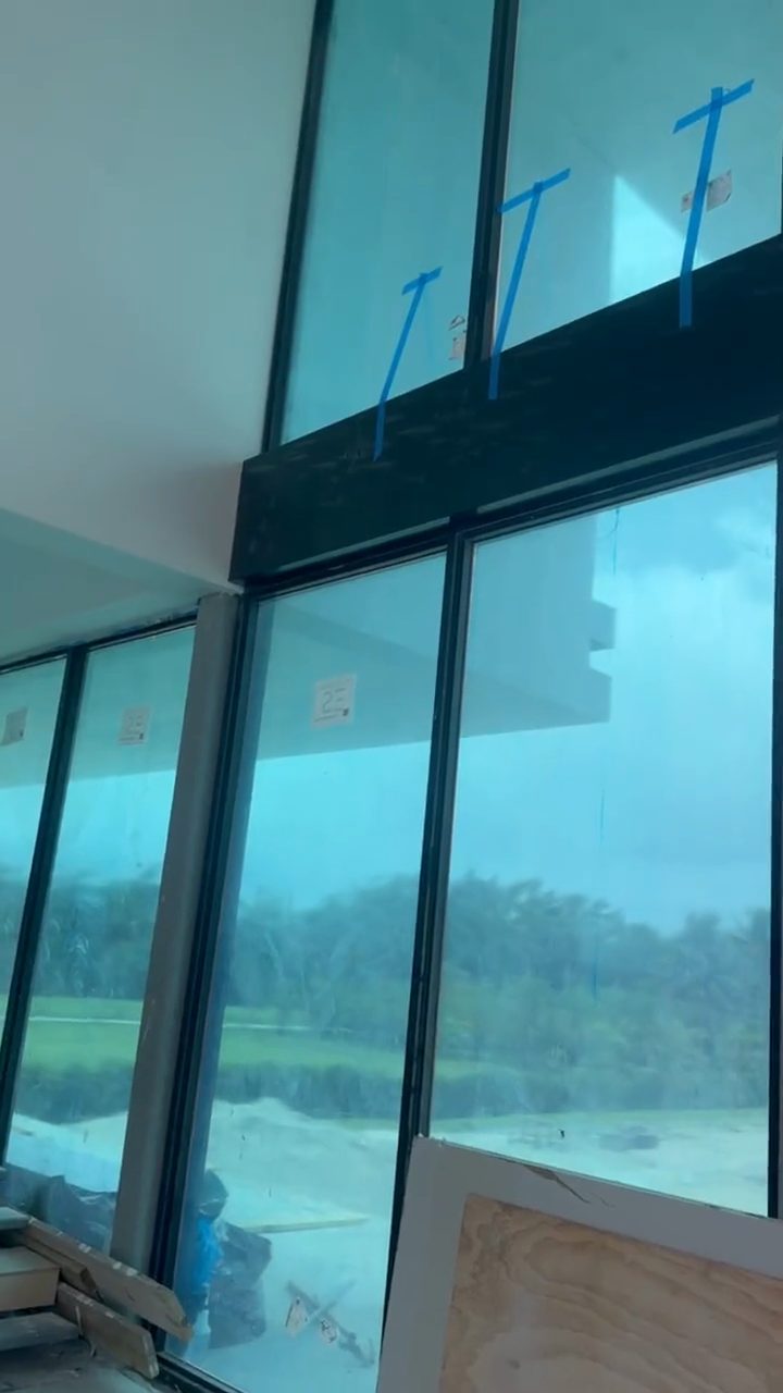 Looking up at a full-height glazed wall being installed inside a large building, the dark frames divided into tall bays, installer tape still crossing the glass and scaffold towers standing against it