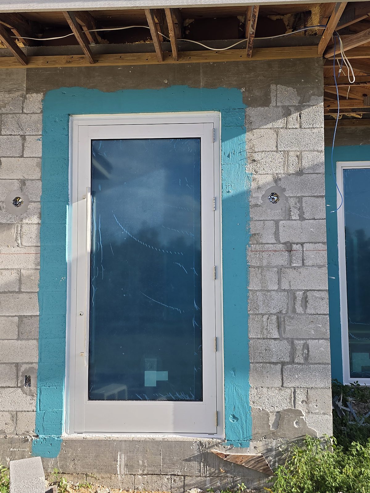 A single white-framed entry door glazed for its full height, hung on three hinges in a teal-painted concrete-block opening, protective blue film still on the glass, on a building still under construction