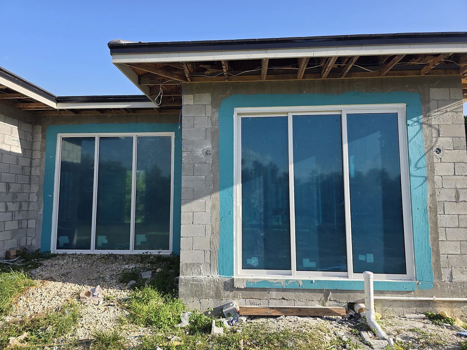 Two openings of new white-framed sliding impact windows in a painted concrete-block wall, protective blue film still on the glass, beneath an unfinished timber eave