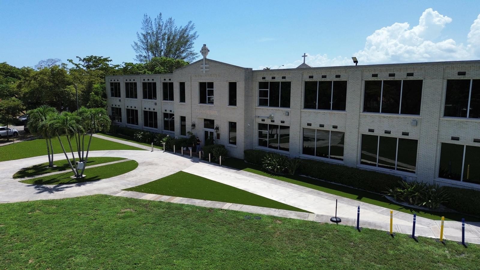 A long two-storey institutional building in pale brick, with rows of dark-framed windows across both floors and full-height glazing along the ground floor, set behind lawns, palms and paved walkways