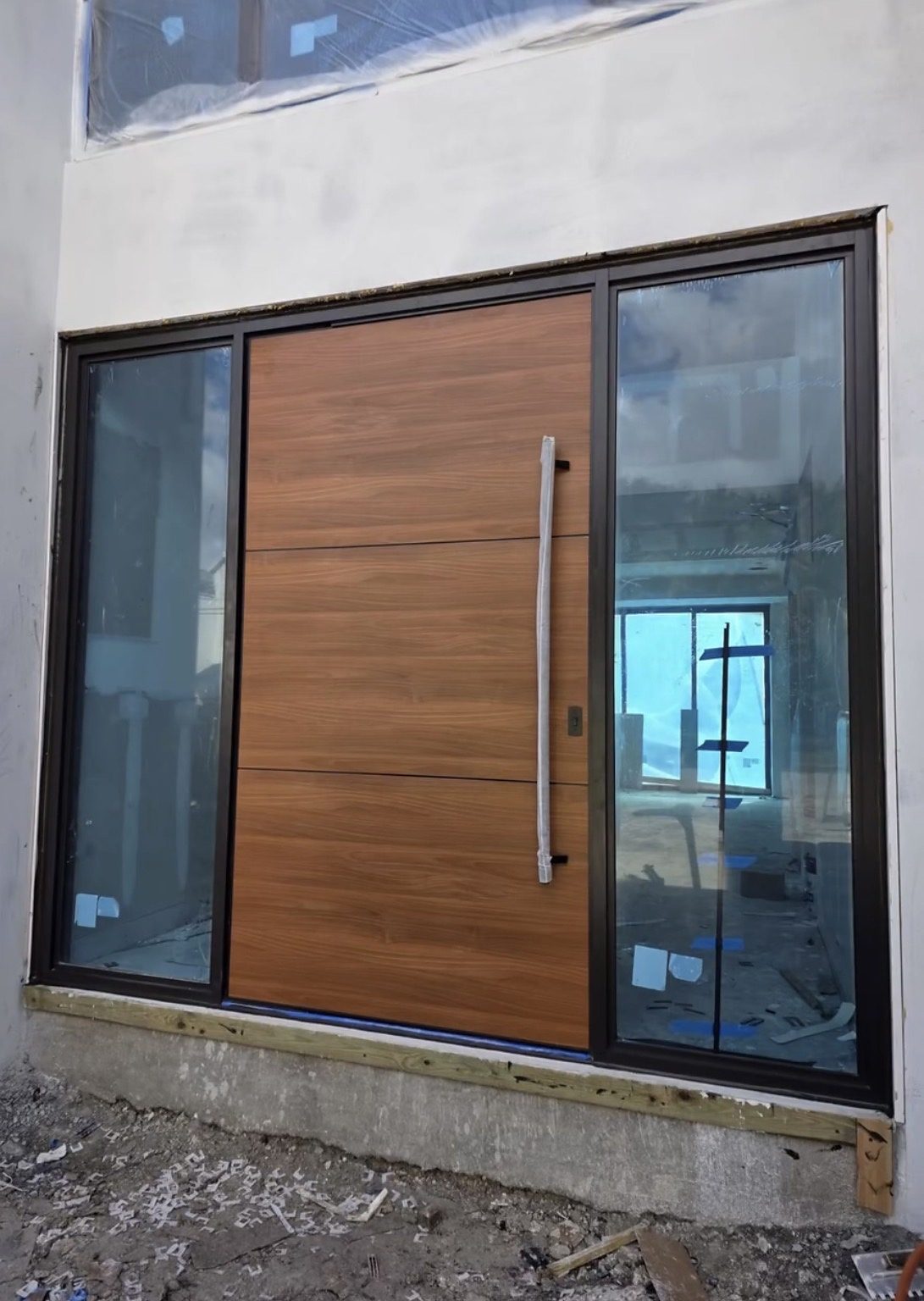 A wood-grain entry door in a dark frame between two tall glazed sidelites, its long pull handle still in a protective wrap, set in a wall still being finished