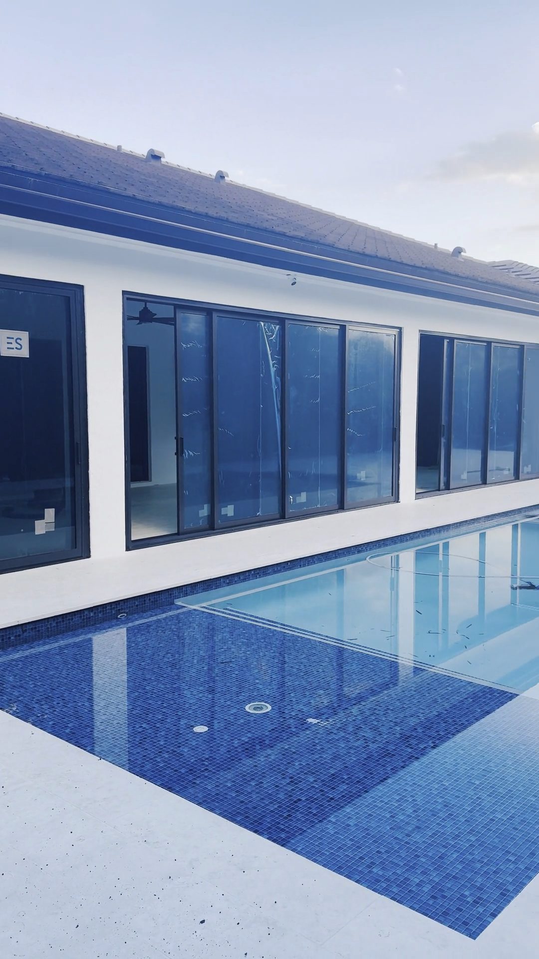 A long run of new dark-framed sliding glass doors along a pool deck, protective labels still on the glass and a ladder standing against the wall