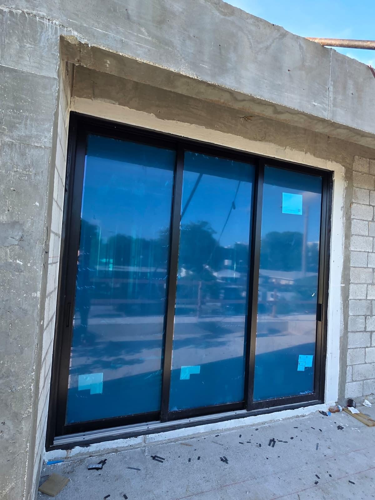 A three-panel bronze-framed sliding glass door set into a concrete-block opening, protective blue film and labels still on the glass, on a building still under construction