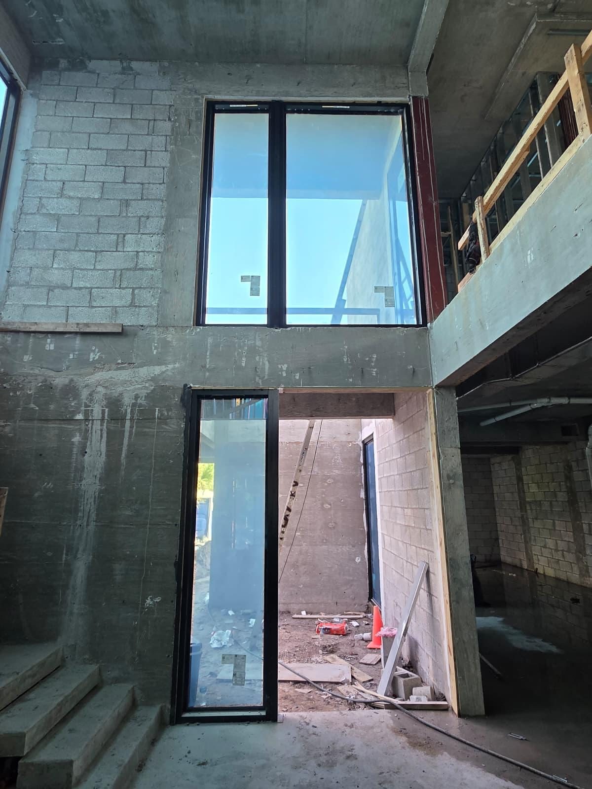 Looking up inside an unfinished concrete and block building at a two-storey stairwell wall, a tall dark-framed glazed opening high in the wall above a full-height fixed glazed panel reaching the floor, with a timber mezzanine alongside