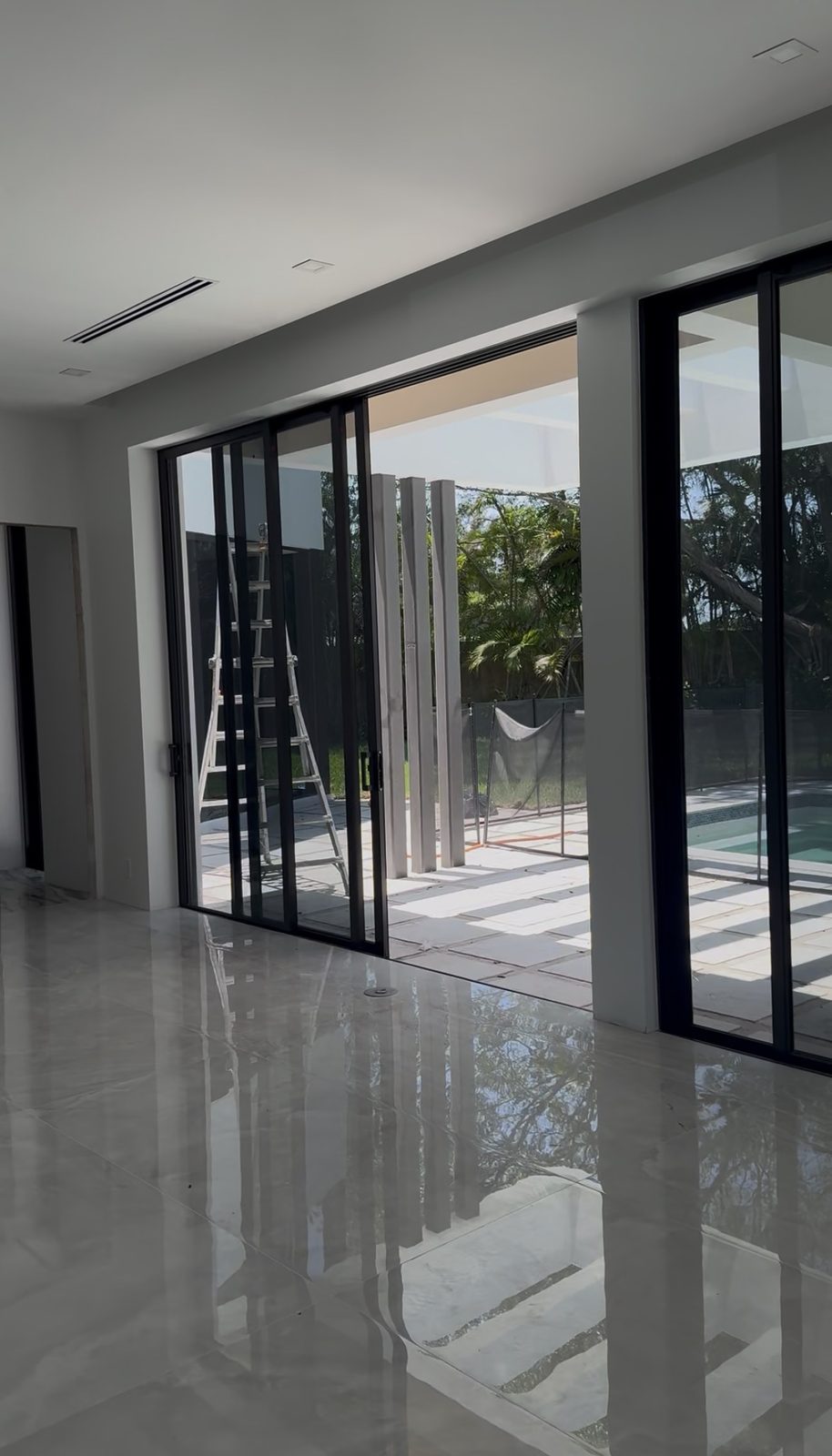 Looking across a polished stone floor through a wall of dark-framed sliding glass doors, standing open onto a covered terrace and a pool beyond