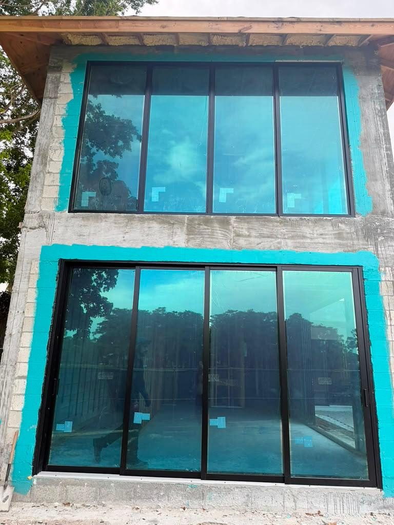 A two-storey glazed elevation on a block building, a four-bay run of dark-framed fixed glass above and a four-panel dark-framed sliding glass door below, teal waterproofing painted around both openings beneath a timber soffit