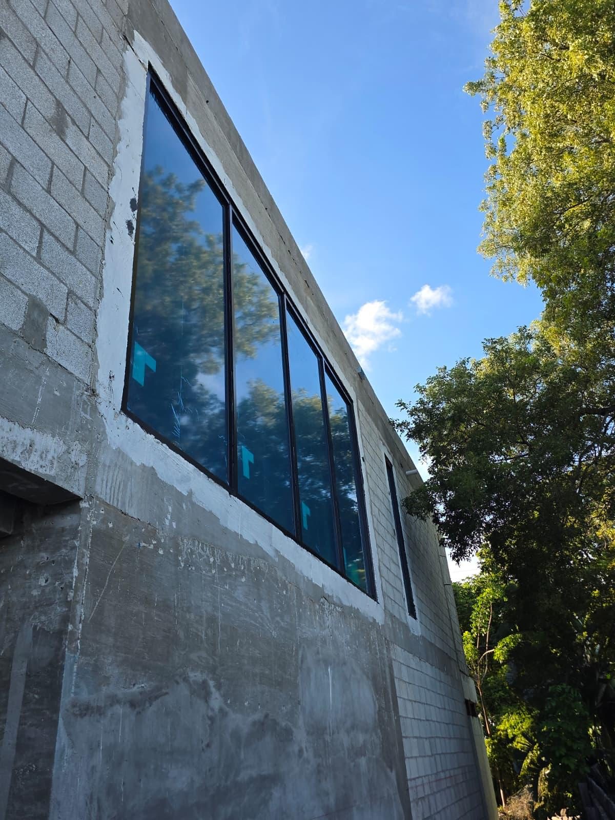 A band of new dark-framed fixed glass set high in a concrete-block wall, seen from below against a blue sky, protective film still on the panes and a tree alongside