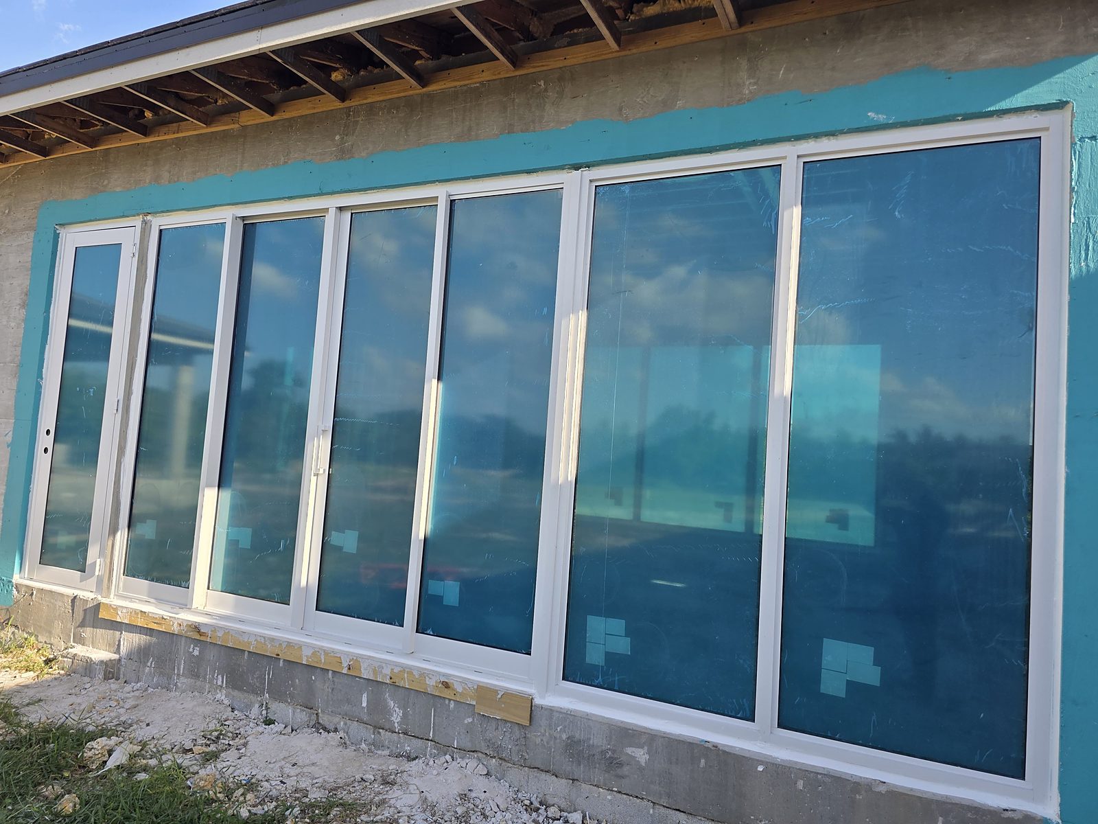 A long run of new white-framed impact glazing filling a wall in seven tall bays, protective blue film still on the panes, with a narrow glazed door at one end, on a block house still under construction
